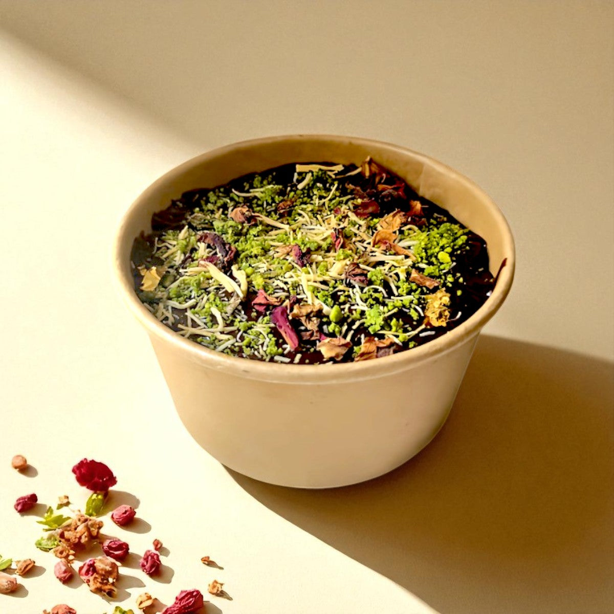 Bowl of chocolate and kunafa surronded by dried roses on a light surface with a soft shadow